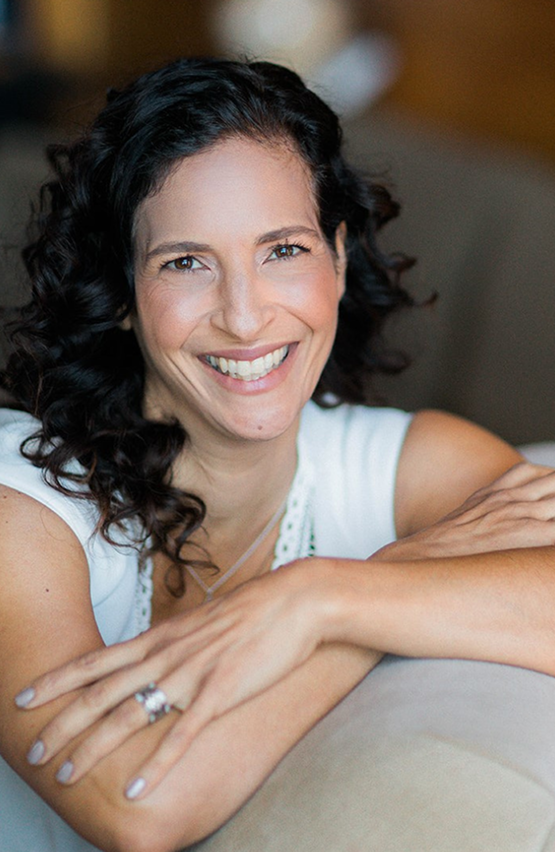 Portrait of a smiling woman with curly dark hair leaning on a cushion in a warmly lit setting.