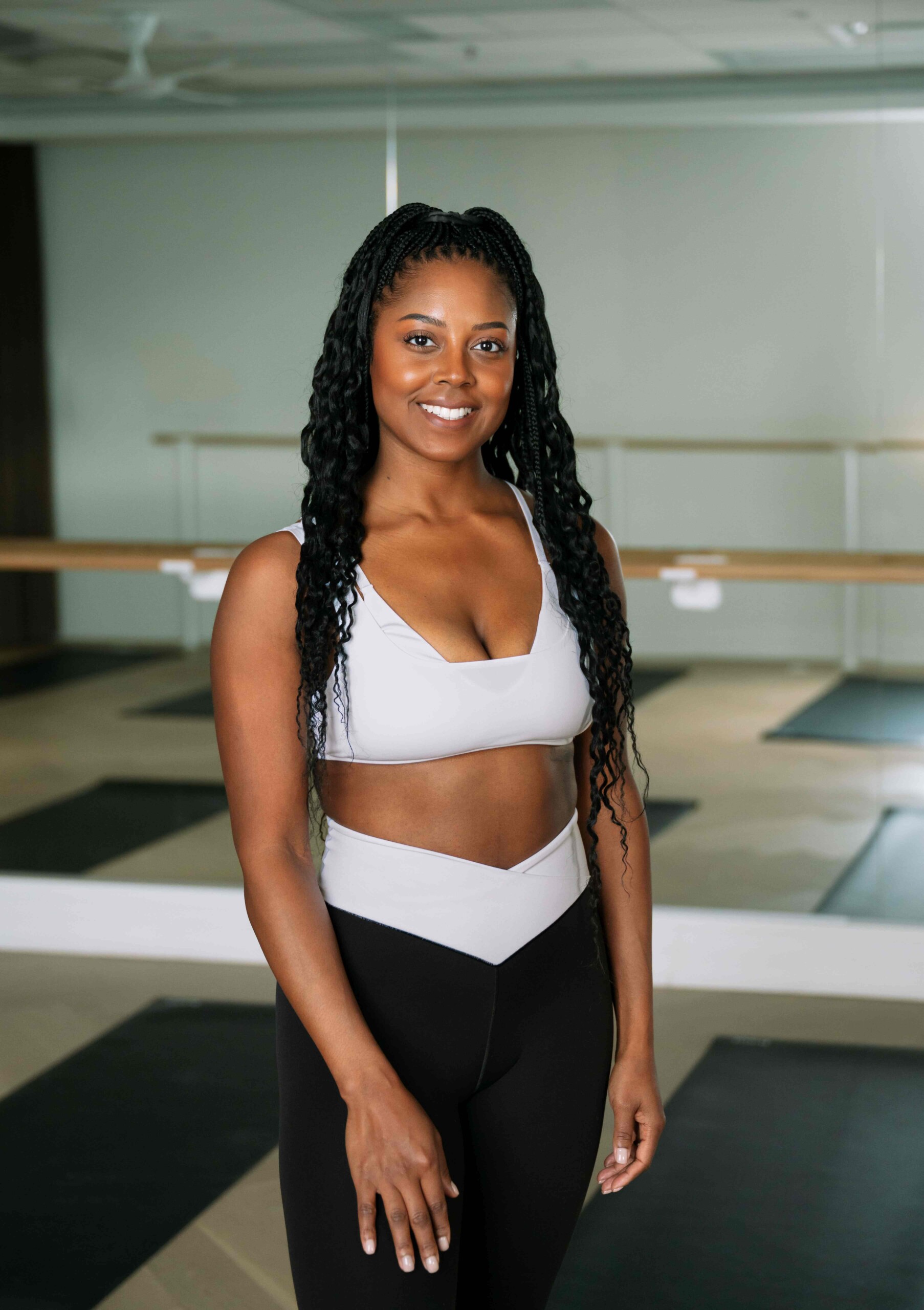 Woman in a light sports bra and black leggings standing in a mirrored yoga studio, smiling at the camera.