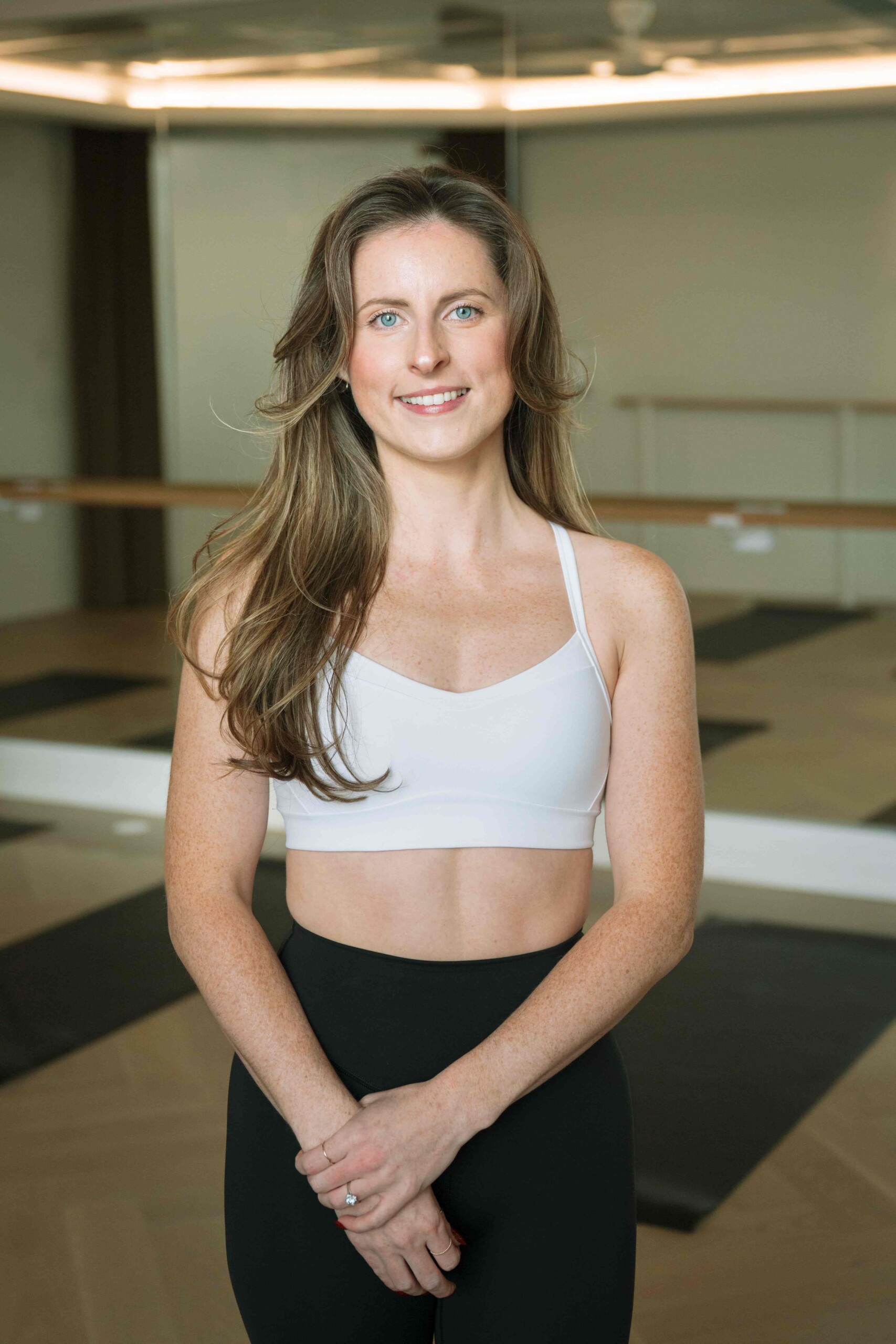 Fitness instructor wearing a light sports bra and black leggings standing in a mirrored studio.