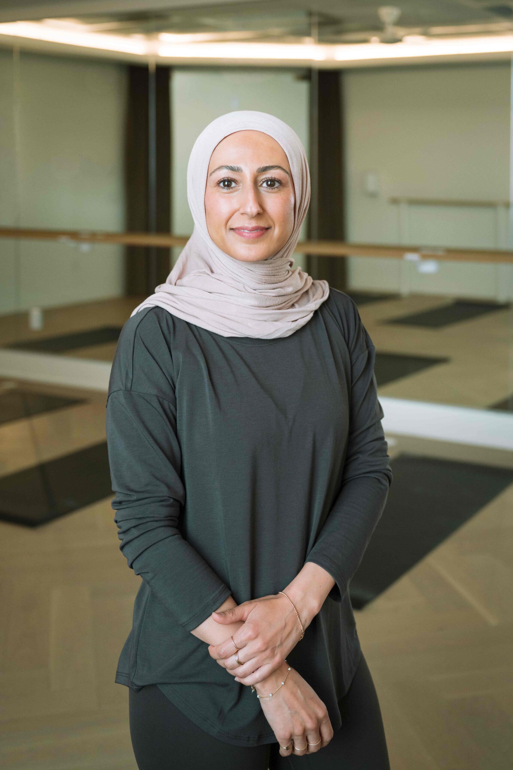 Woman wearing a light hijab and dark long-sleeve top standing in a mirrored yoga studio.