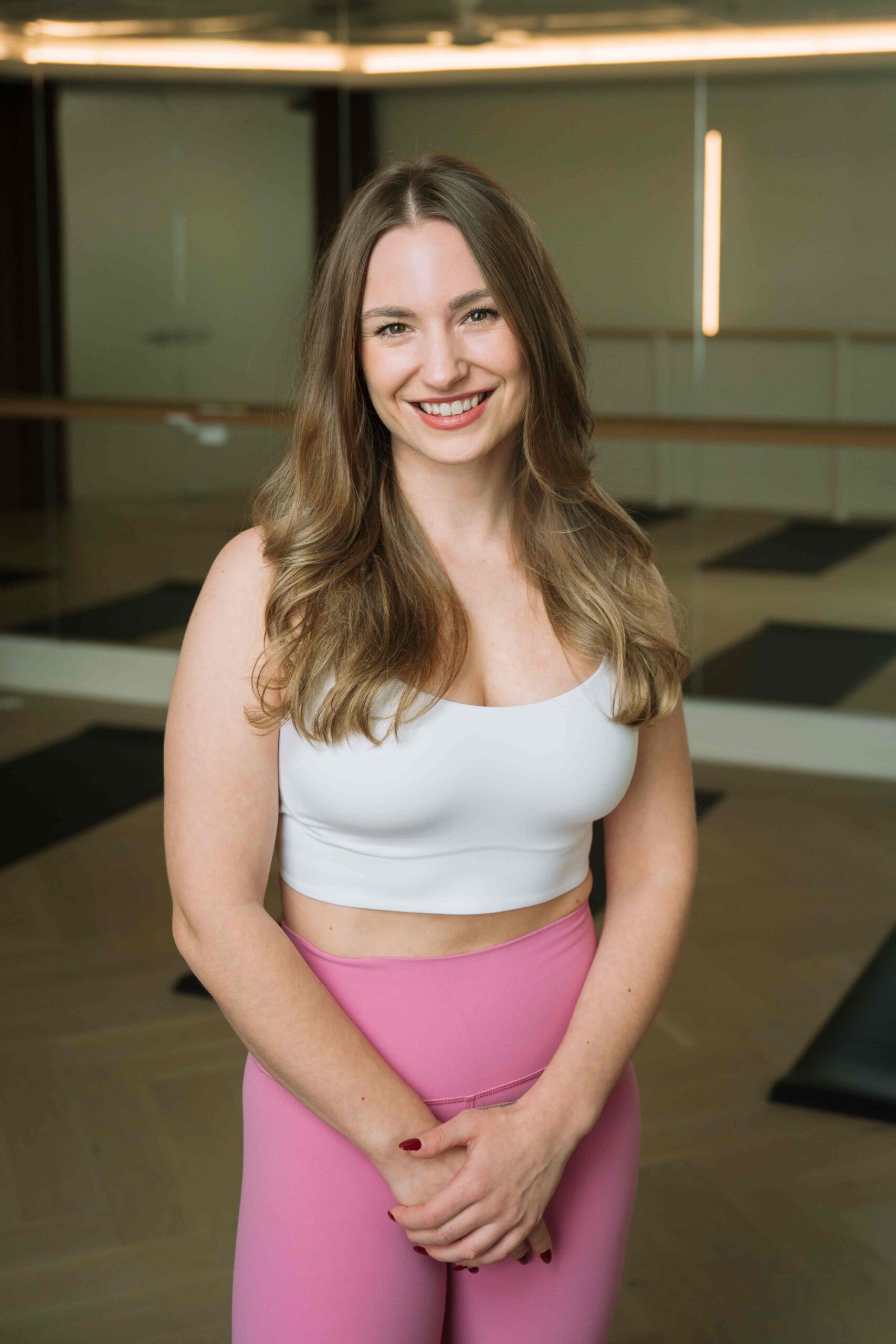 Woman in a light sports bra and pink leggings standing in a mirrored fitness studio, smiling at the camera.