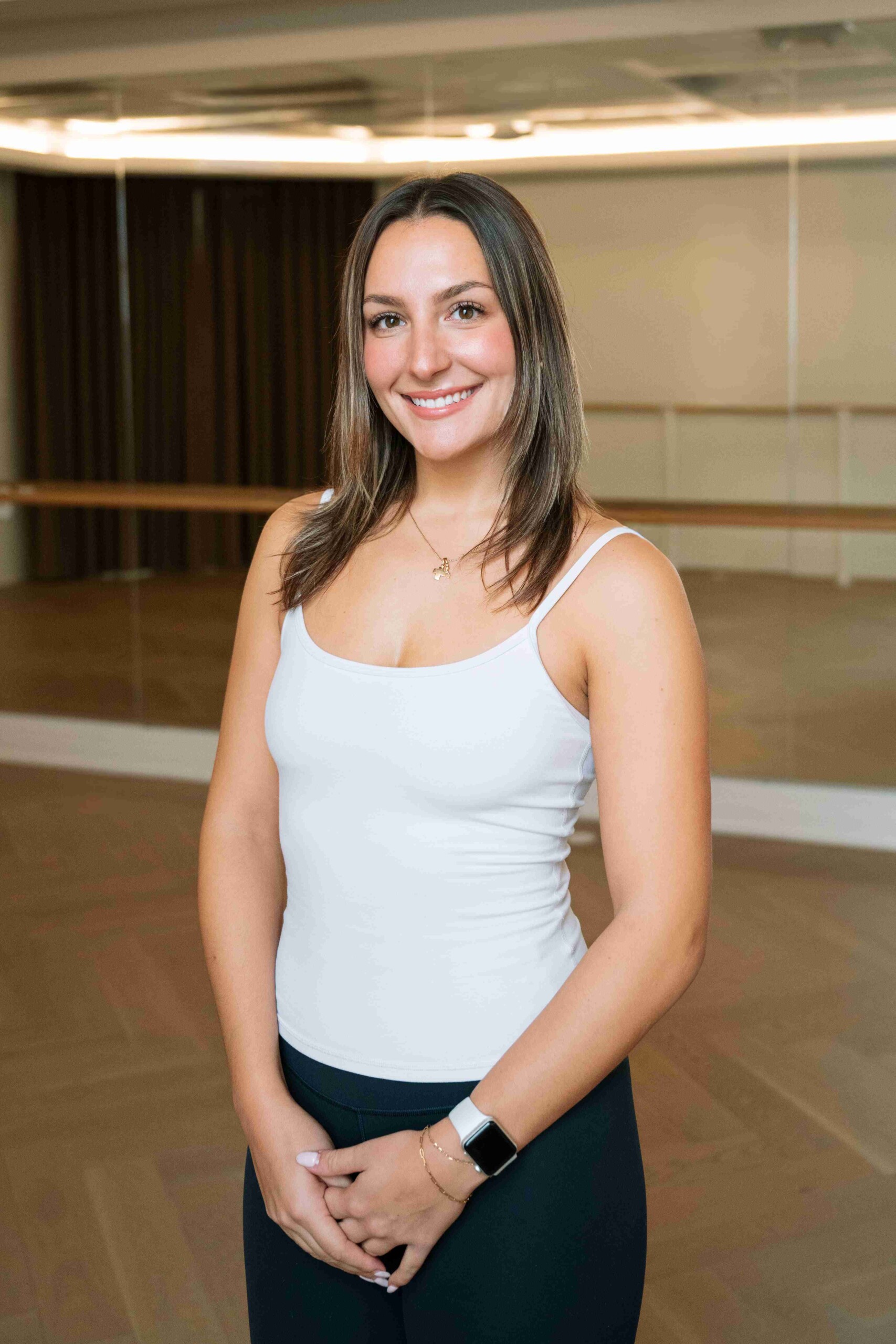 Woman in a light tank top and black leggings standing in a mirrored fitness studio, smiling at the camera.