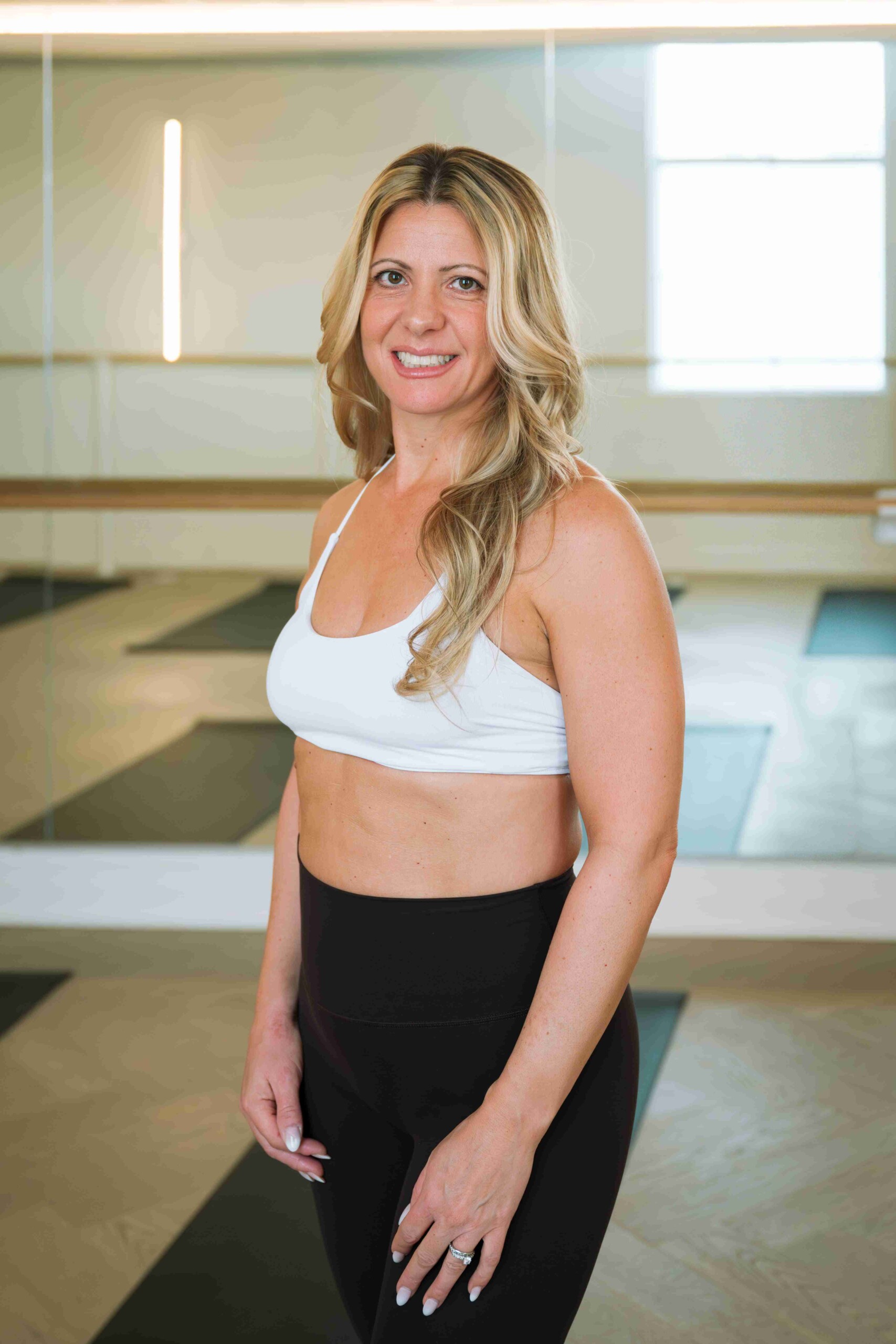 Woman in a white sports bra and black leggings standing in a yoga studio with mirrors and mats in the background.